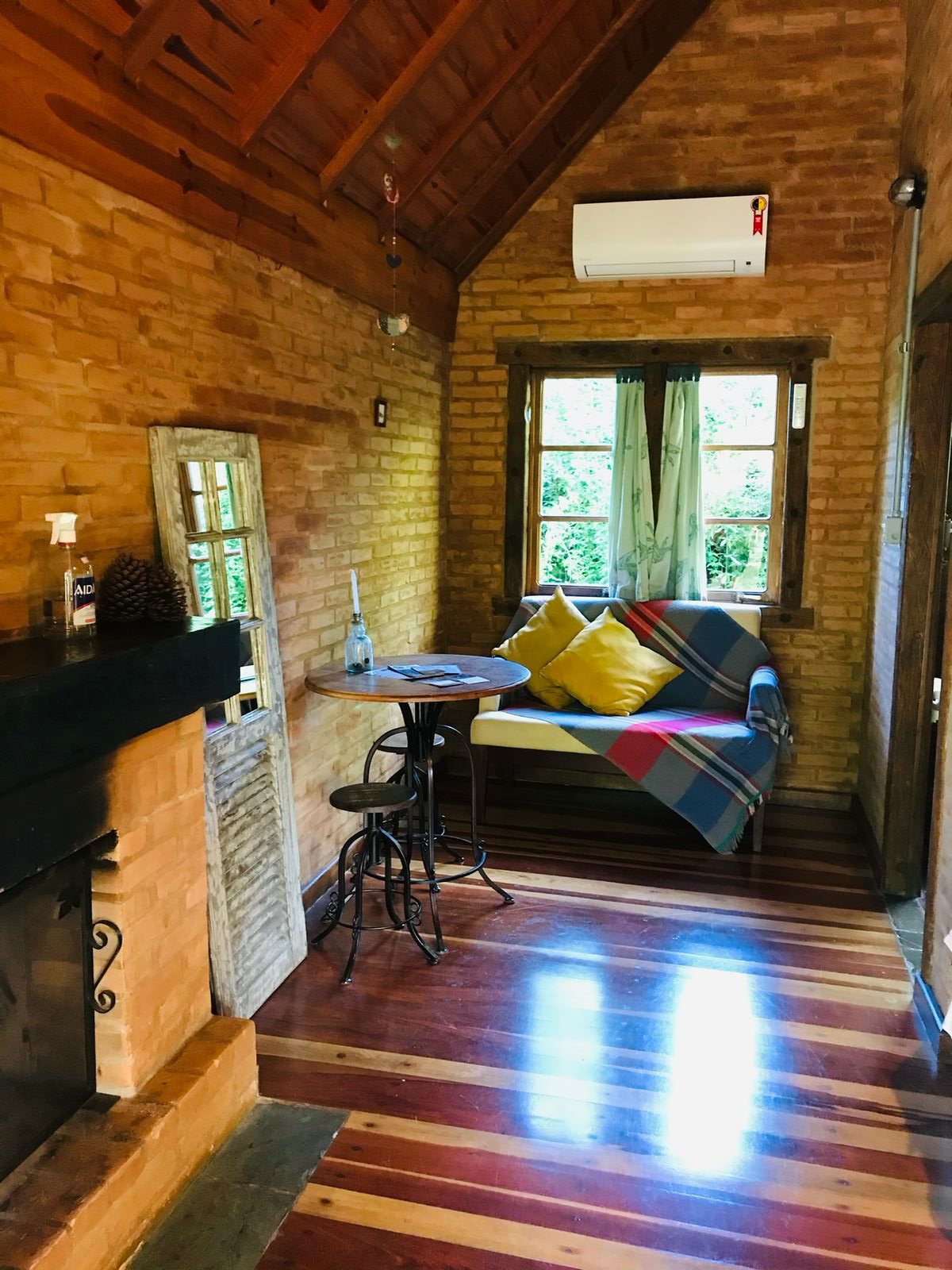 Sala do Chalé Ipê: lareira, mesa de ferro com banquetas e sofá com almofadas amarelas junto à janela