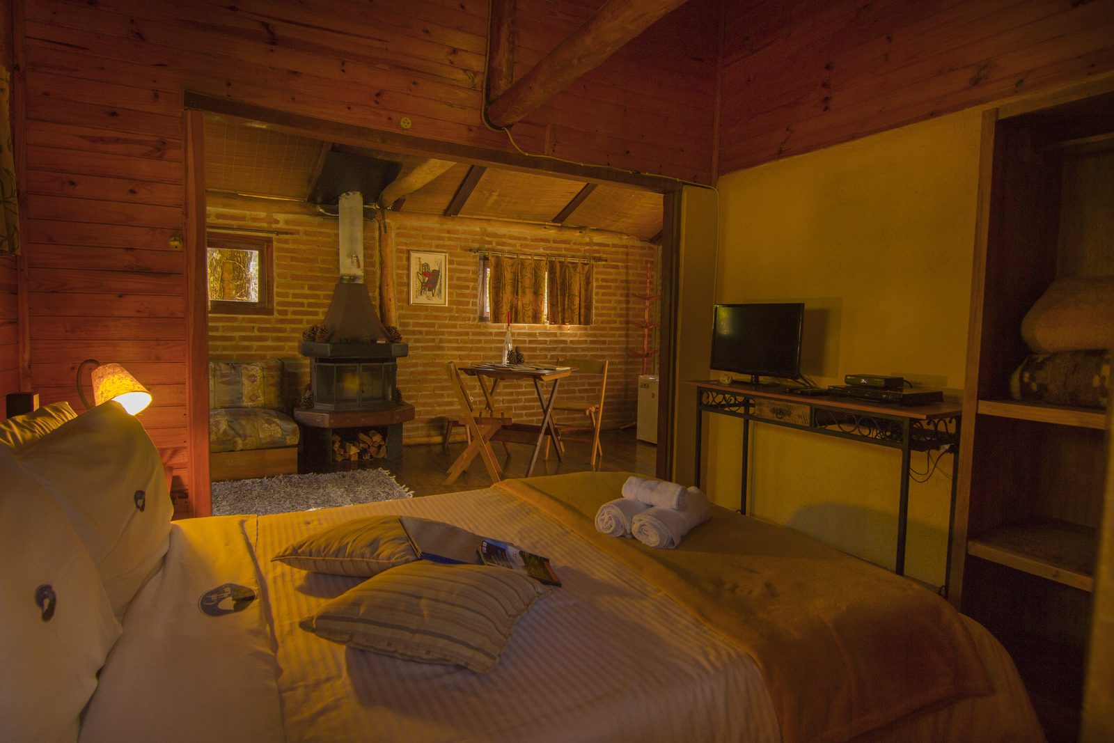 Quarto do Chalé Paineiras com a cama, o abajur aceso e a sala com o fogão a lenha ao fundo