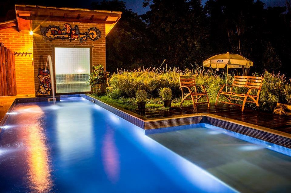 Piscina iluminada à noite, com a sauna de vidro ao fundo
